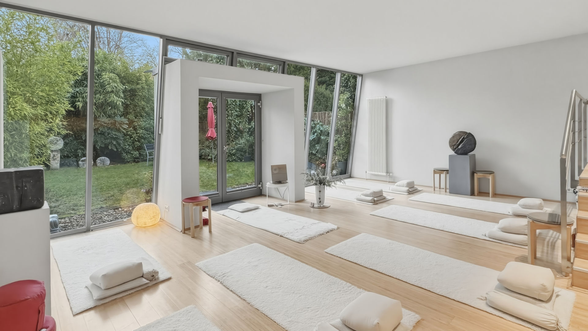 Innenansicht des Yogastudios mit ausgelegten Matten, Meditationskissen und großen schrägen Fensterfronten mit Blick nach draußen.